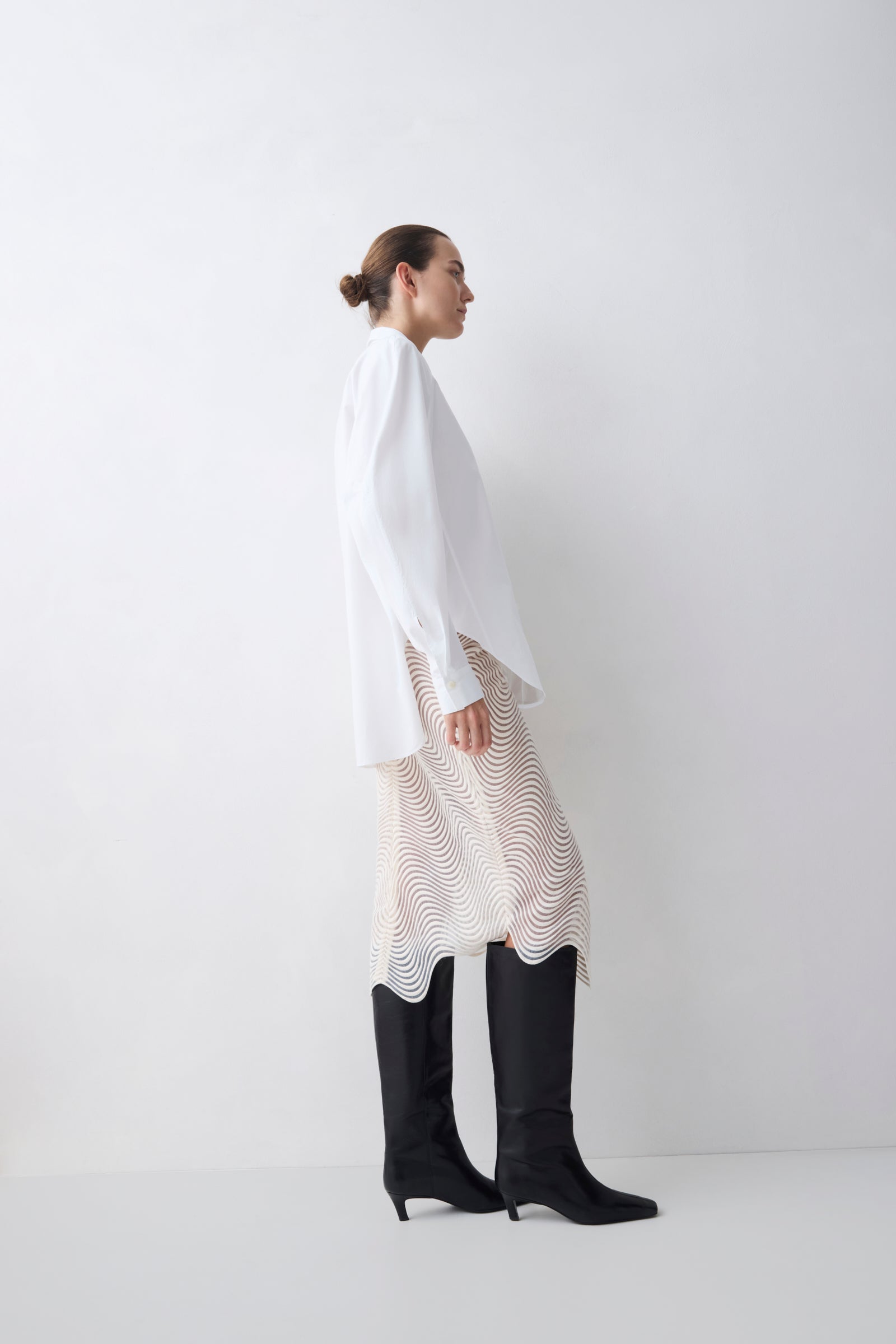 Person wearing a white long-sleeve top, patterned skirt, and black knee-high boots on a white background