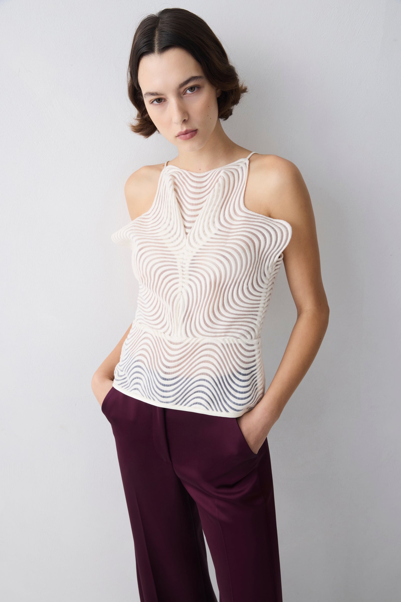Woman wearing a white sleeveless top with a wavy pattern and purple pants against a light gray background