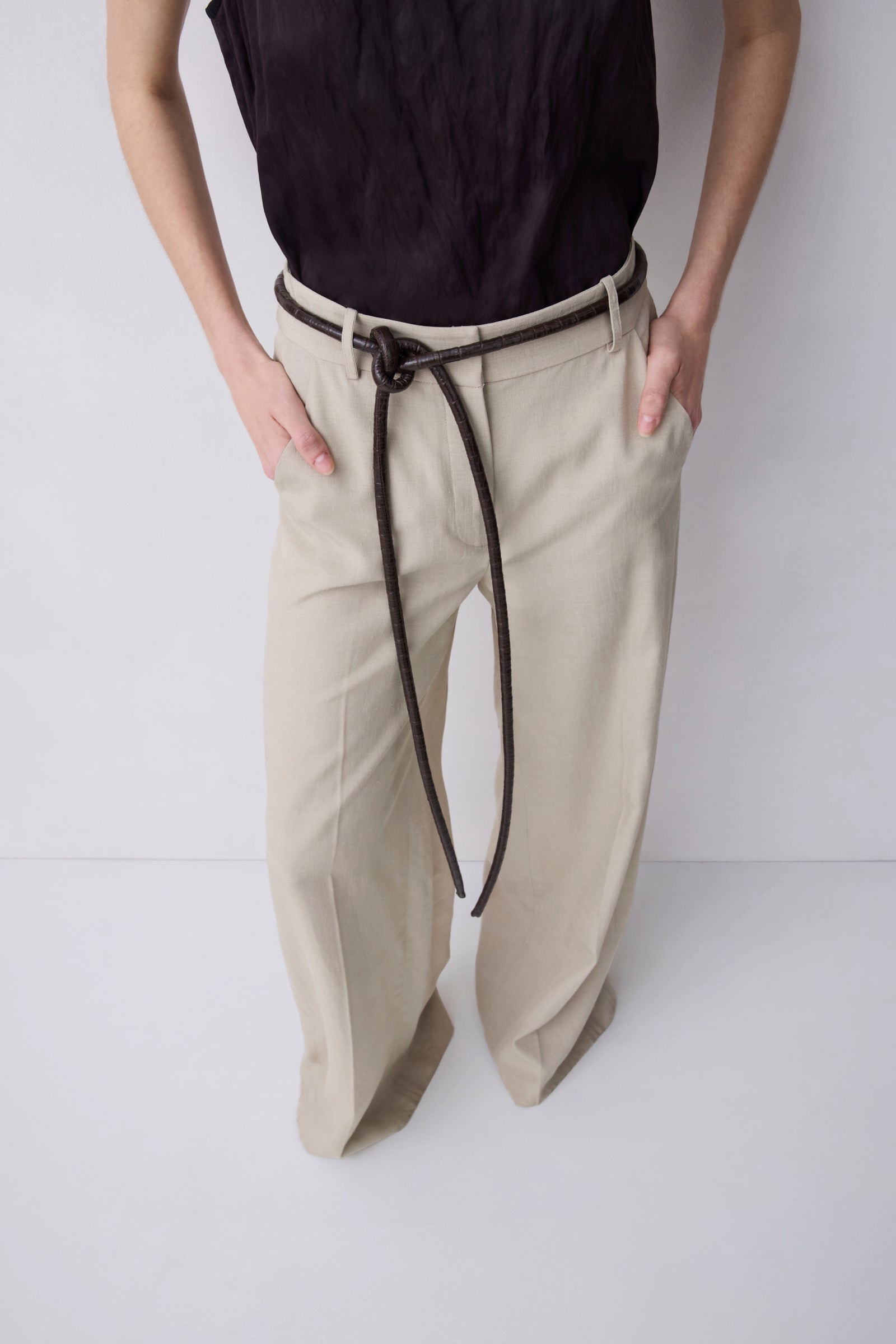 A person wearing beige wide-leg trousers with a dark brown rope belt tied at the waist, paired with a black sleeveless top, against a plain light gray studio background.