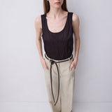 A woman stands against a plain light gray studio background wearing a sleeveless black top and loose beige pants with a black drawstring.