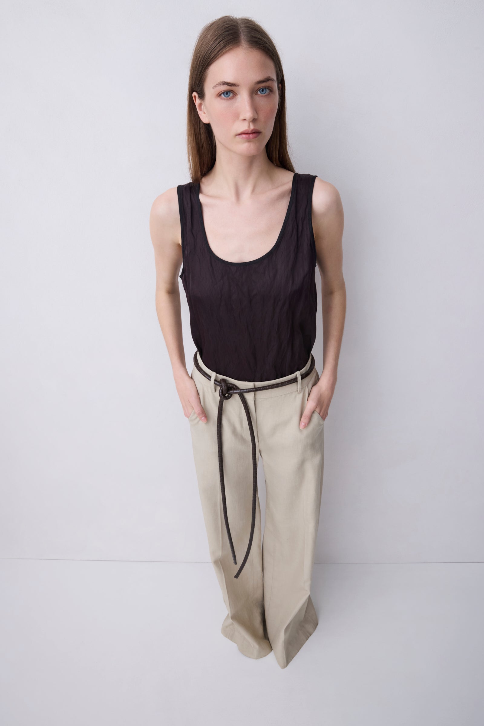 A woman stands against a plain light gray studio background wearing a sleeveless black top and loose beige pants with a black drawstring.