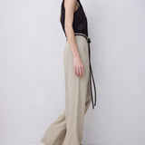 A woman stands in profile wearing a black sleeveless top and high-waisted beige wide-leg trousers with a thin black belt, photographed against a plain light gray studio background.
