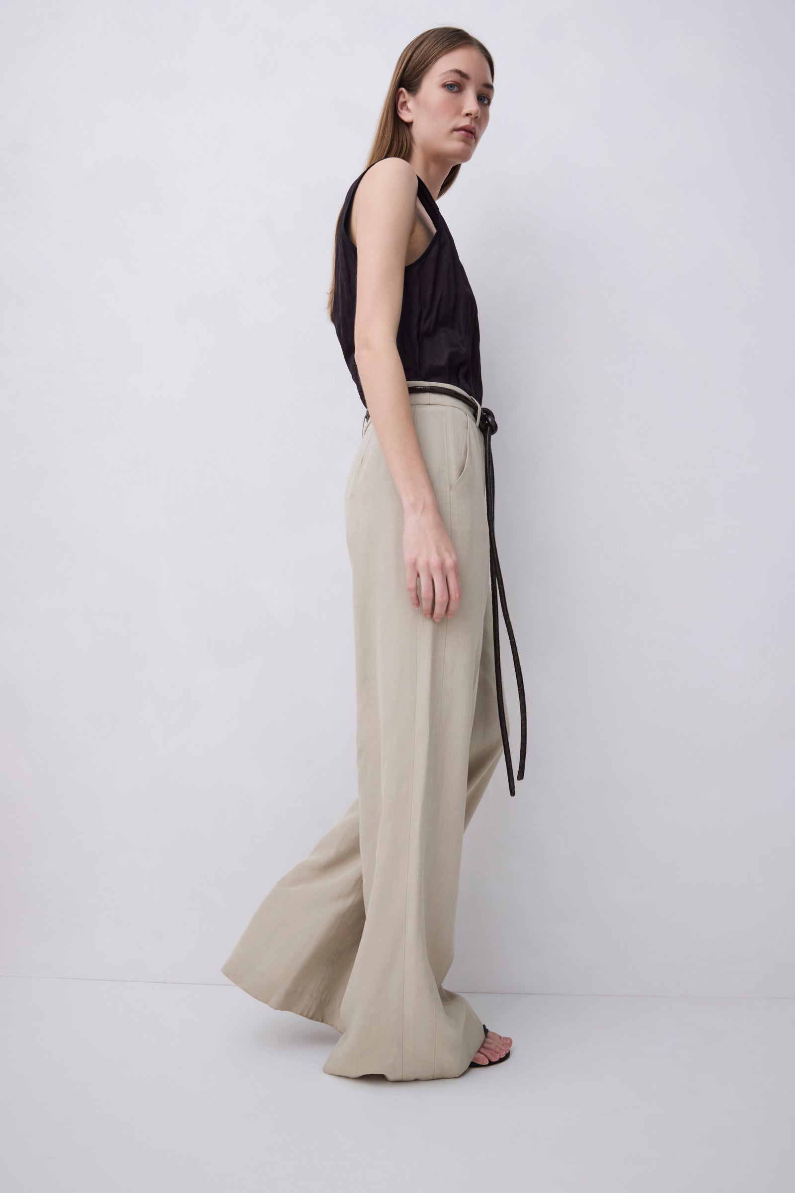 A woman stands in profile wearing a black sleeveless top and high-waisted beige wide-leg trousers with a thin black belt, photographed against a plain light gray studio background.
