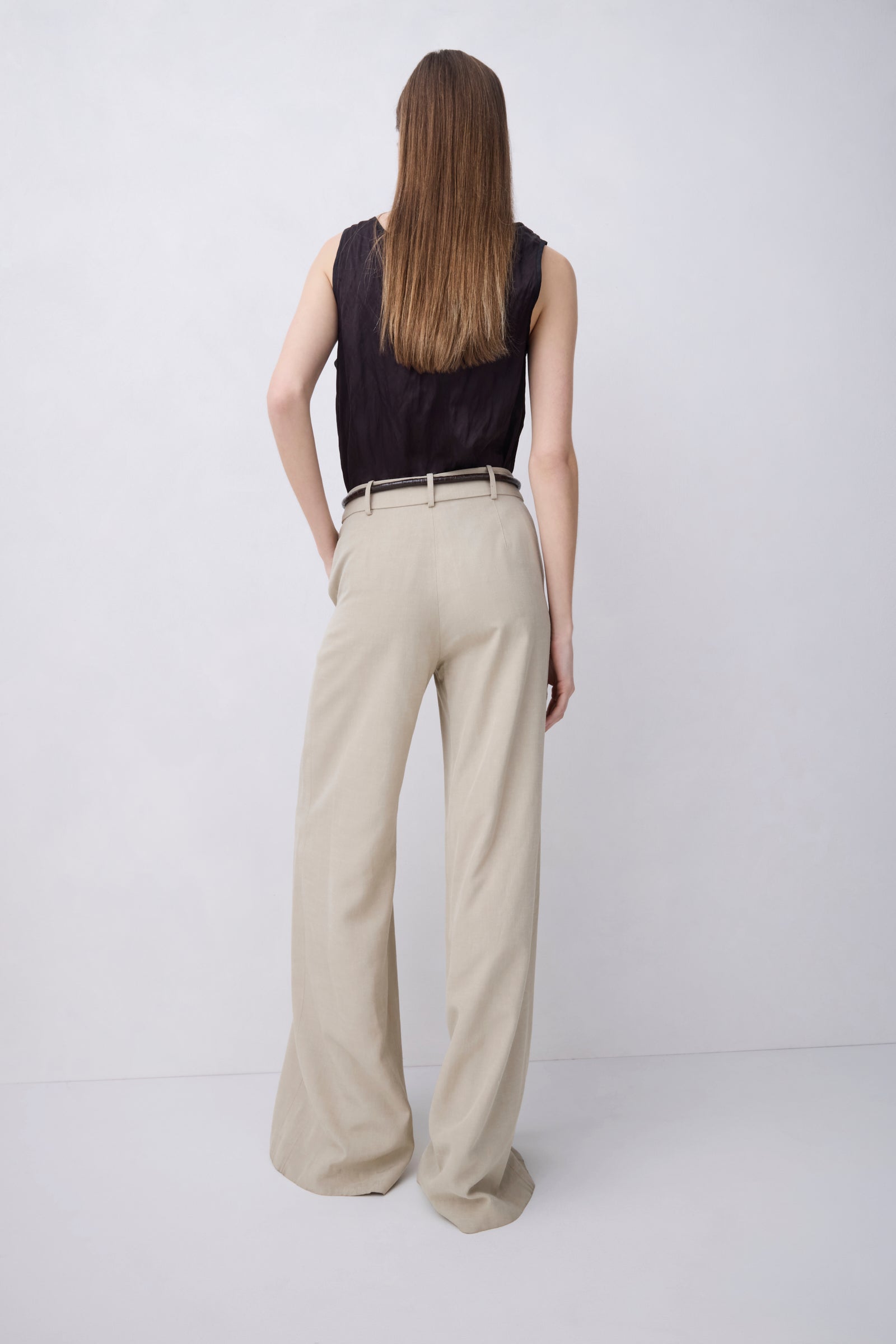 A woman is shown from behind wearing a black sleeveless top and beige wide-leg trousers with a thin black belt, standing against a light gray studio background.