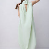 A woman viewed from behind wearing a flowing pale mint green dress with a large draped neckline and open back, standing against a plain light gray studio background.