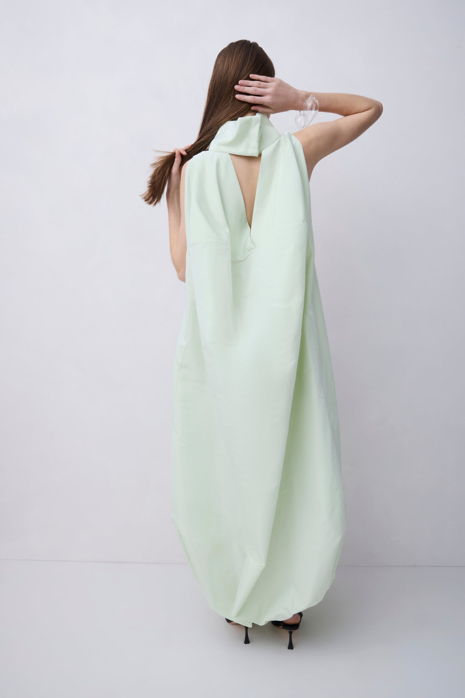 A woman viewed from behind wearing a flowing pale mint green dress with a large draped neckline and open back, standing against a plain light gray studio background.