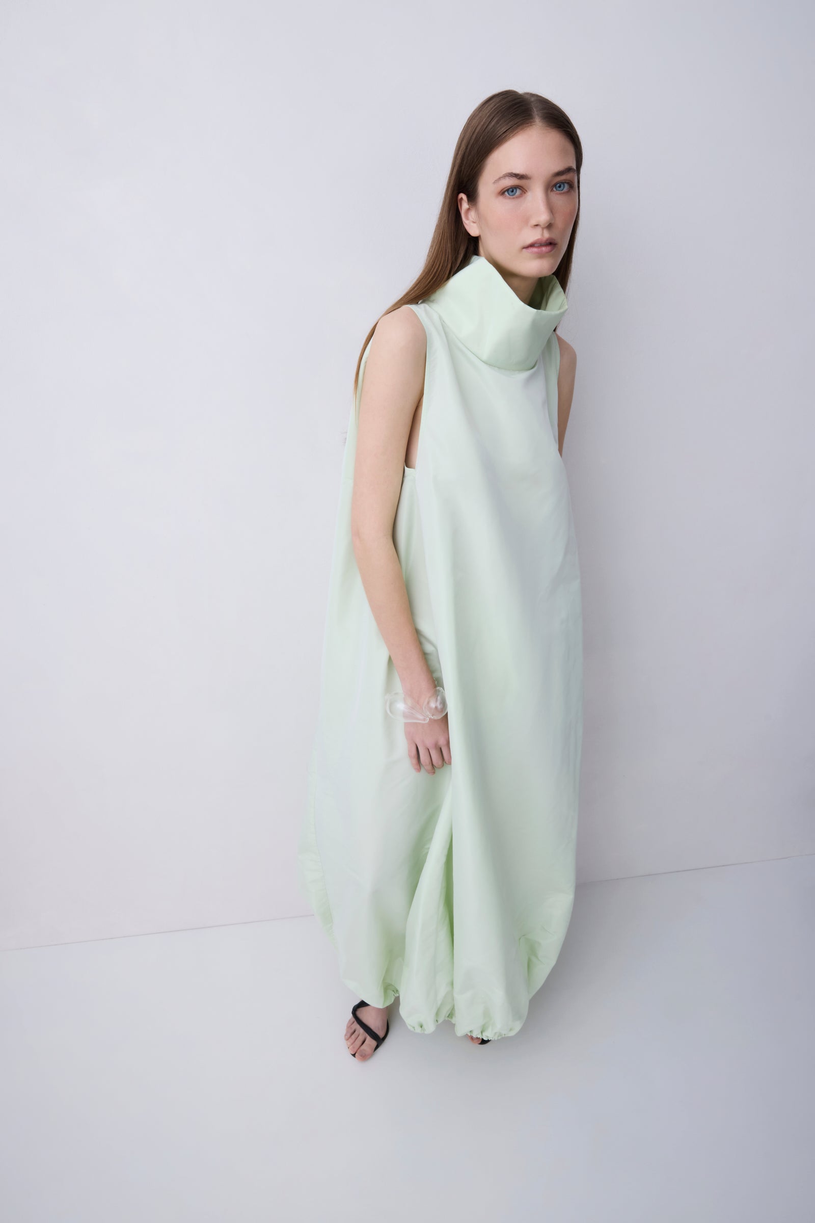 A woman modeling a sleeveless, high-neck, flowing maxi dress in a pale mint green color, standing against a plain light gray studio background.