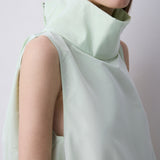A close-up of a model wearing a pale mint green sleeveless top with a high structured collar and draped fabric detail, set against a plain light gray background.