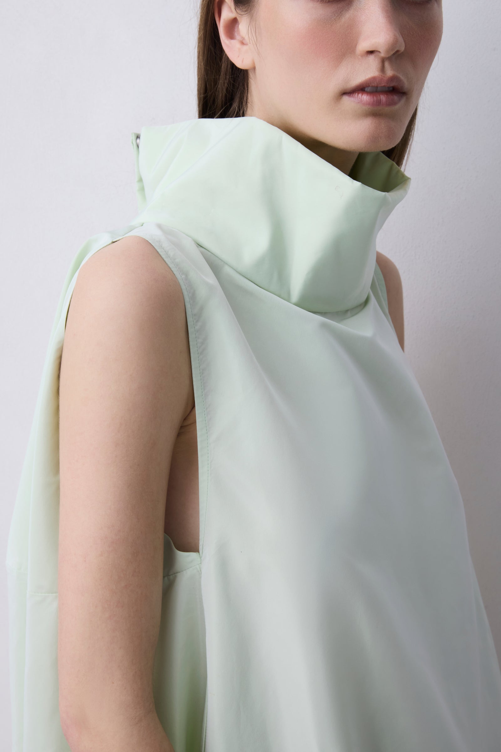 A close-up of a model wearing a pale mint green sleeveless top with a high structured collar and draped fabric detail, set against a plain light gray background.