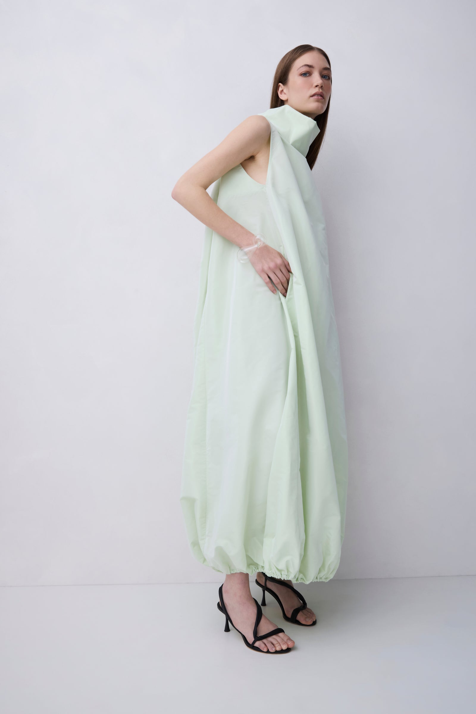 A model wearing a pale mint green sleeveless maxi dress with a high draped collar and voluminous gathered hem, paired with black strappy high-heeled sandals, against a plain light gray studio background.