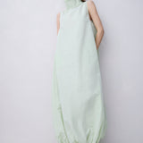 A model wearing a pale mint green sleeveless maxi dress with a high mock neck and gathered hem, paired with black strappy heels, against a plain light gray studio background.