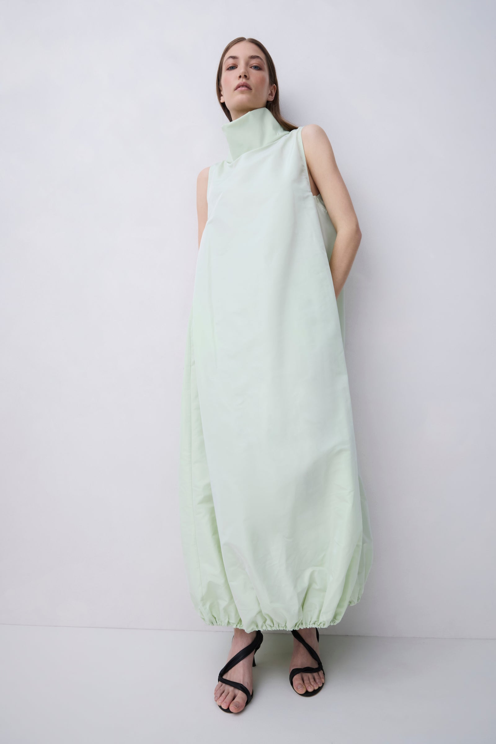A model wearing a pale mint green sleeveless maxi dress with a high mock neck and gathered hem, paired with black strappy heels, against a plain light gray studio background.