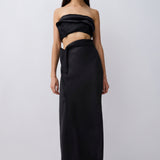 A model wearing a black strapless draped tube top and matching black wrap-style maxi skirt, paired with black pointed flats, against a light gray studio background.
