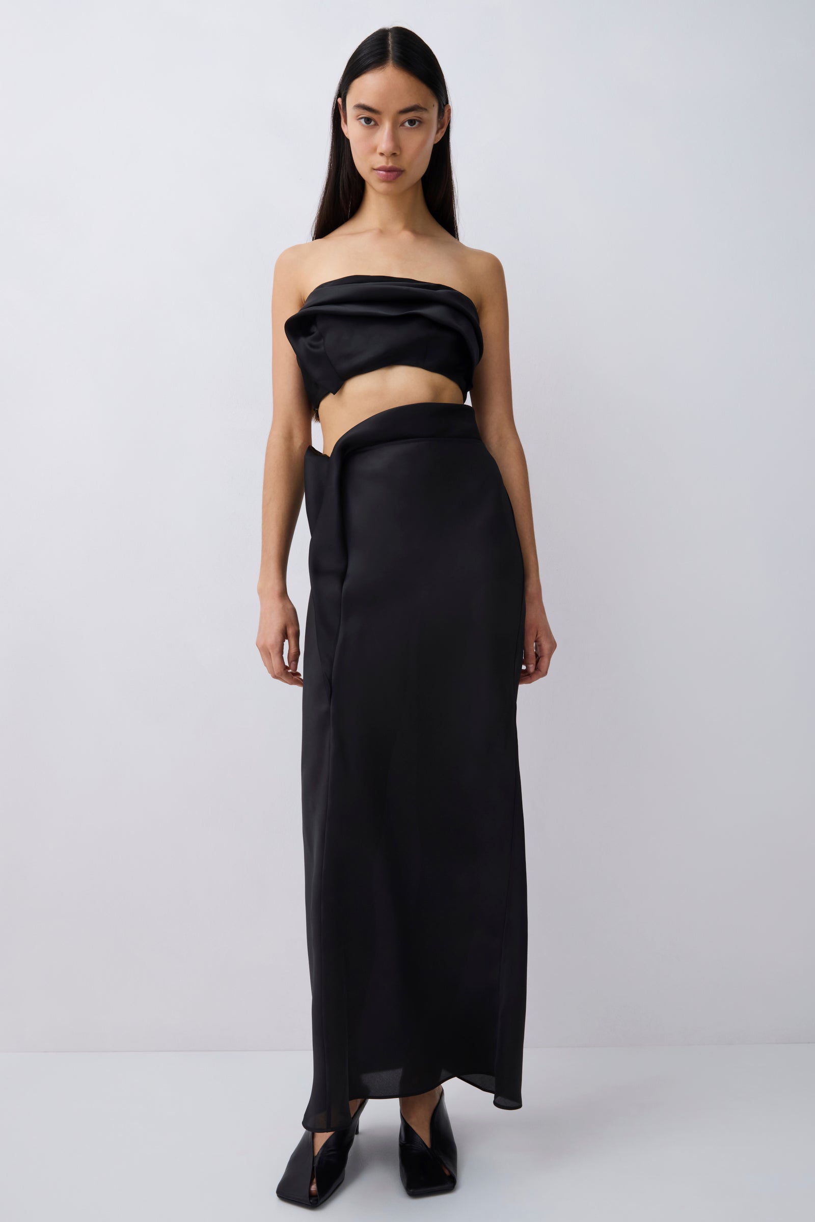 A model wearing a black strapless draped tube top and matching black wrap-style maxi skirt, paired with black pointed flats, against a light gray studio background.