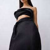 A model wearing a black strapless draped dress with a high-waisted, voluminous skirt against a light gray studio background.