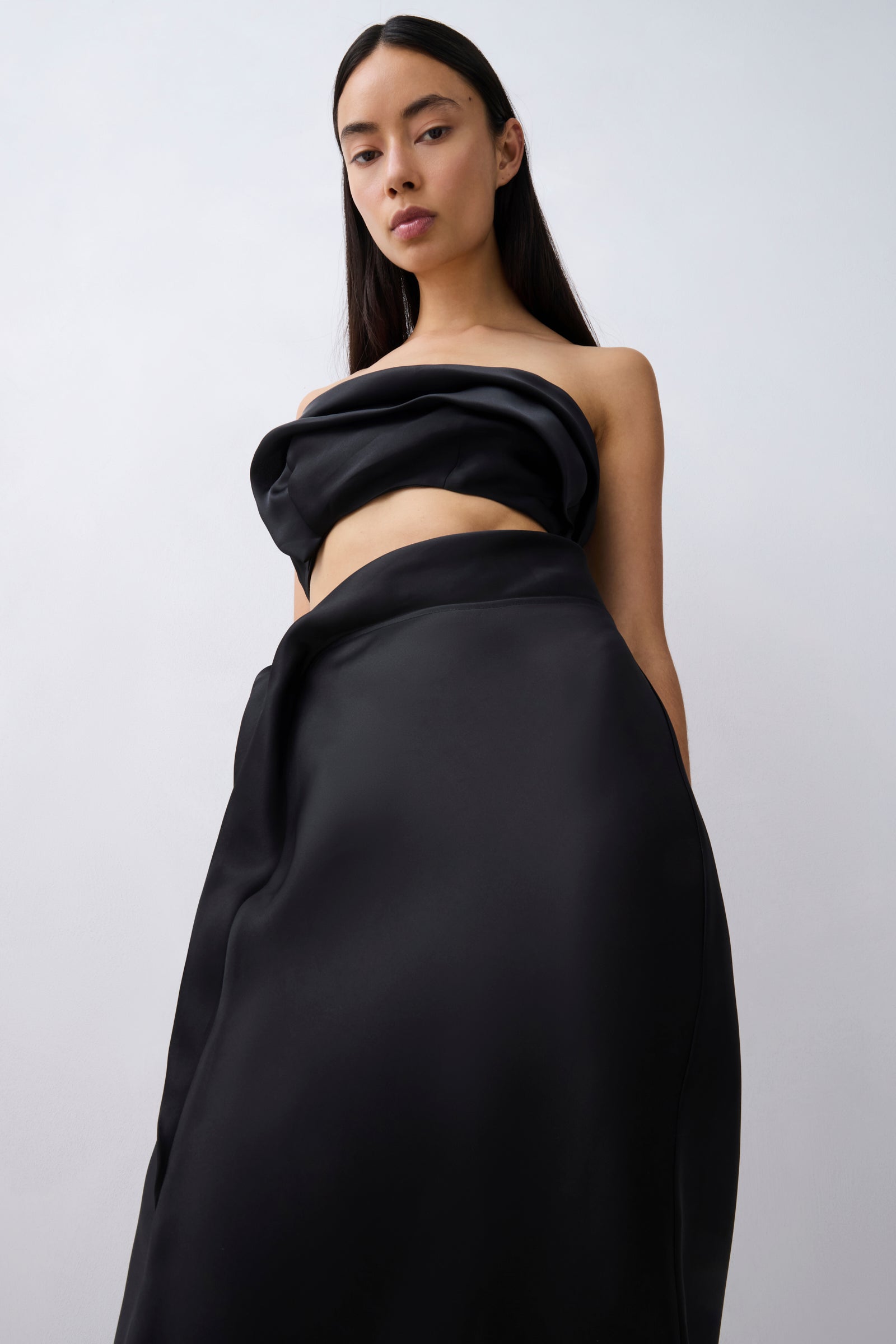 A model wearing a black strapless draped dress with a high-waisted, voluminous skirt against a light gray studio background.