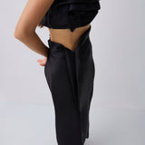 A model shown from the back wearing a black strapless draped crop top and matching black satin-like wide-leg skirt or pants against a light gray studio background.