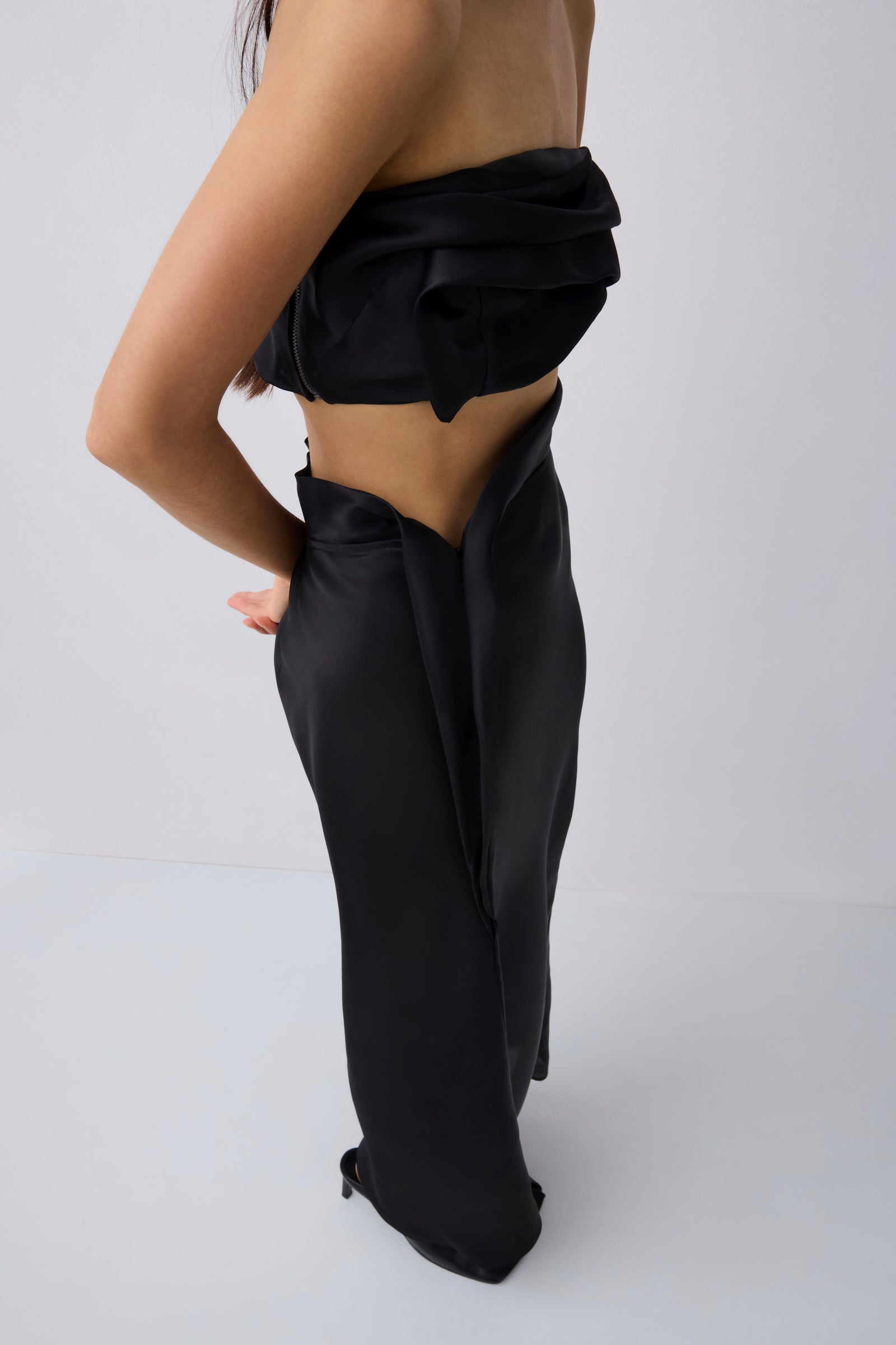 A model shown from the back wearing a black strapless draped crop top and matching black satin-like wide-leg skirt or pants against a light gray studio background.
