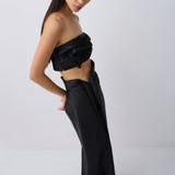 A model wearing a black strapless draped crop top and matching wide-leg draped trousers, styled with black pointed-toe heels against a light gray studio background.