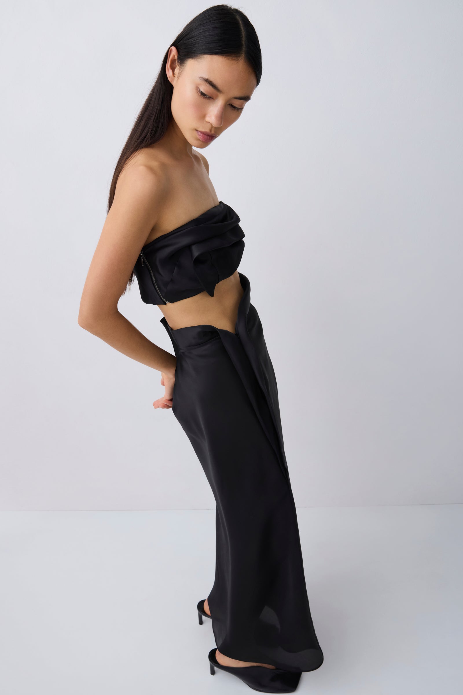 A model wearing a black strapless draped crop top and matching wide-leg draped trousers, styled with black pointed-toe heels against a light gray studio background.