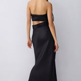 A woman shown from the back wearing a black strapless maxi skirt with a gathered waist, paired with black pointed heels, against a light gray studio background.