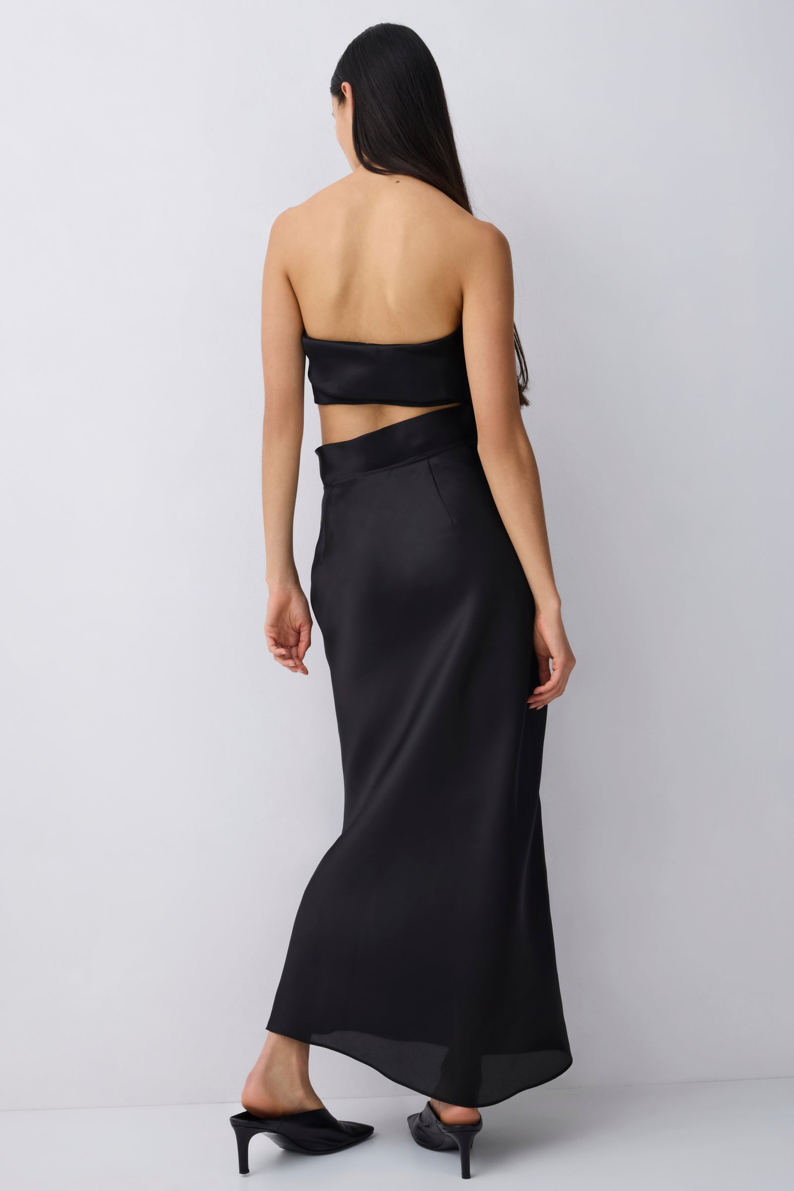 A woman shown from the back wearing a black strapless maxi skirt with a gathered waist, paired with black pointed heels, against a light gray studio background.