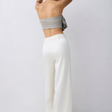 A woman shown from behind wearing a light gray strapless bandeau-style top and high-waisted, wide-leg white trousers against a plain light gray studio background.