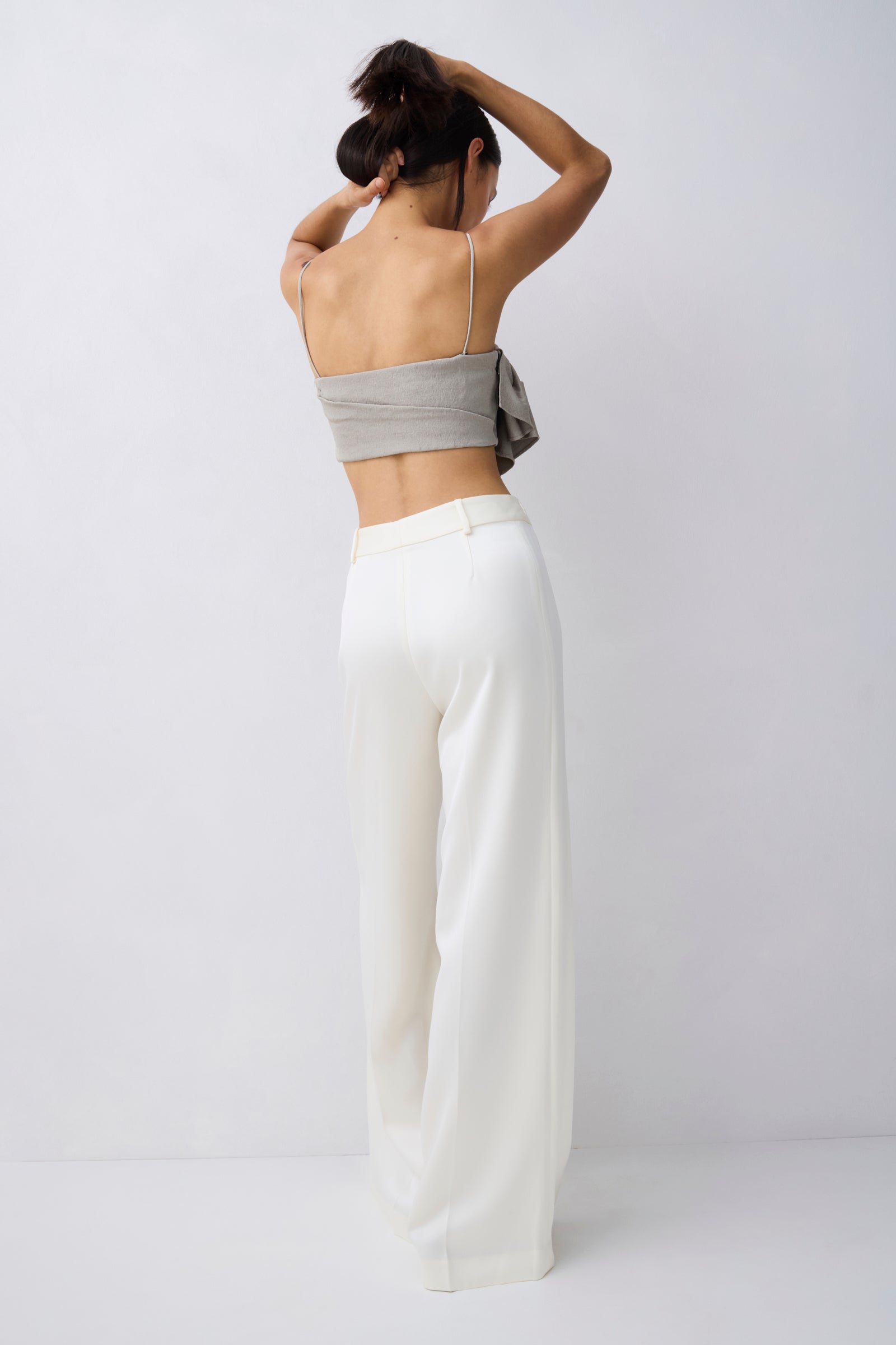A woman shown from behind wearing a light gray strapless bandeau-style top and high-waisted, wide-leg white trousers against a plain light gray studio background.