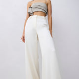 A model wearing a light gray ruffled strapless crop top and high-waisted white wide-leg trousers, standing against a plain light gray studio background.