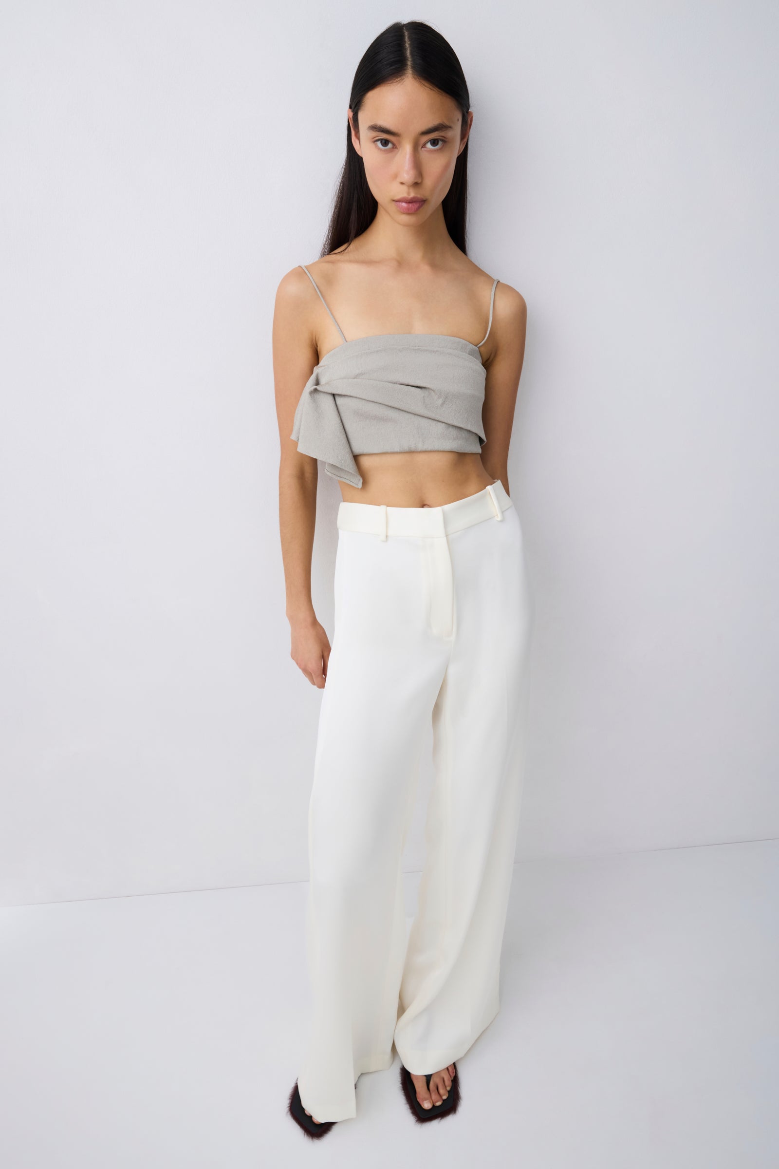 A model stands against a light gray studio wall wearing a light gray wrap-style bandeau top and high-waisted, wide-leg white trousers.