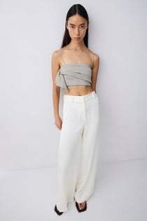 A model stands against a light gray studio wall wearing a light gray wrap-style bandeau top and high-waisted, wide-leg white trousers.