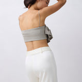 A woman shown from the back wearing a light gray bandeau-style crop top with thin straps and a large side bow, paired with white high-waisted pants, against a plain light gray studio background.