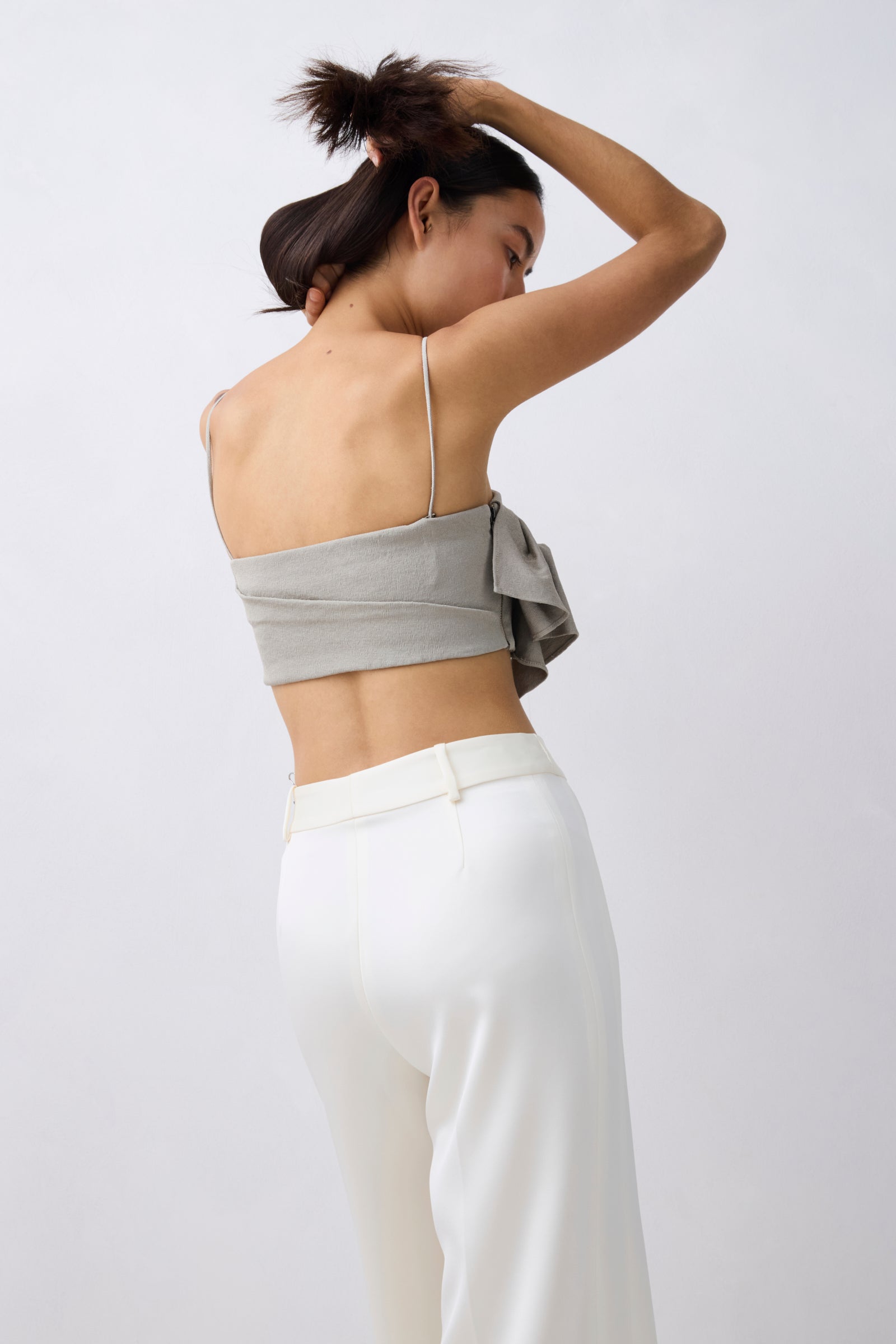 A woman shown from the back wearing a light gray bandeau-style crop top with thin straps and a large side bow, paired with white high-waisted pants, against a plain light gray studio background.