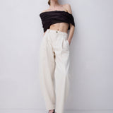 A model stands against a light gray studio wall wearing a black off-the-shoulder cropped top, cream wide-leg trousers, and black strappy high-heeled sandals.