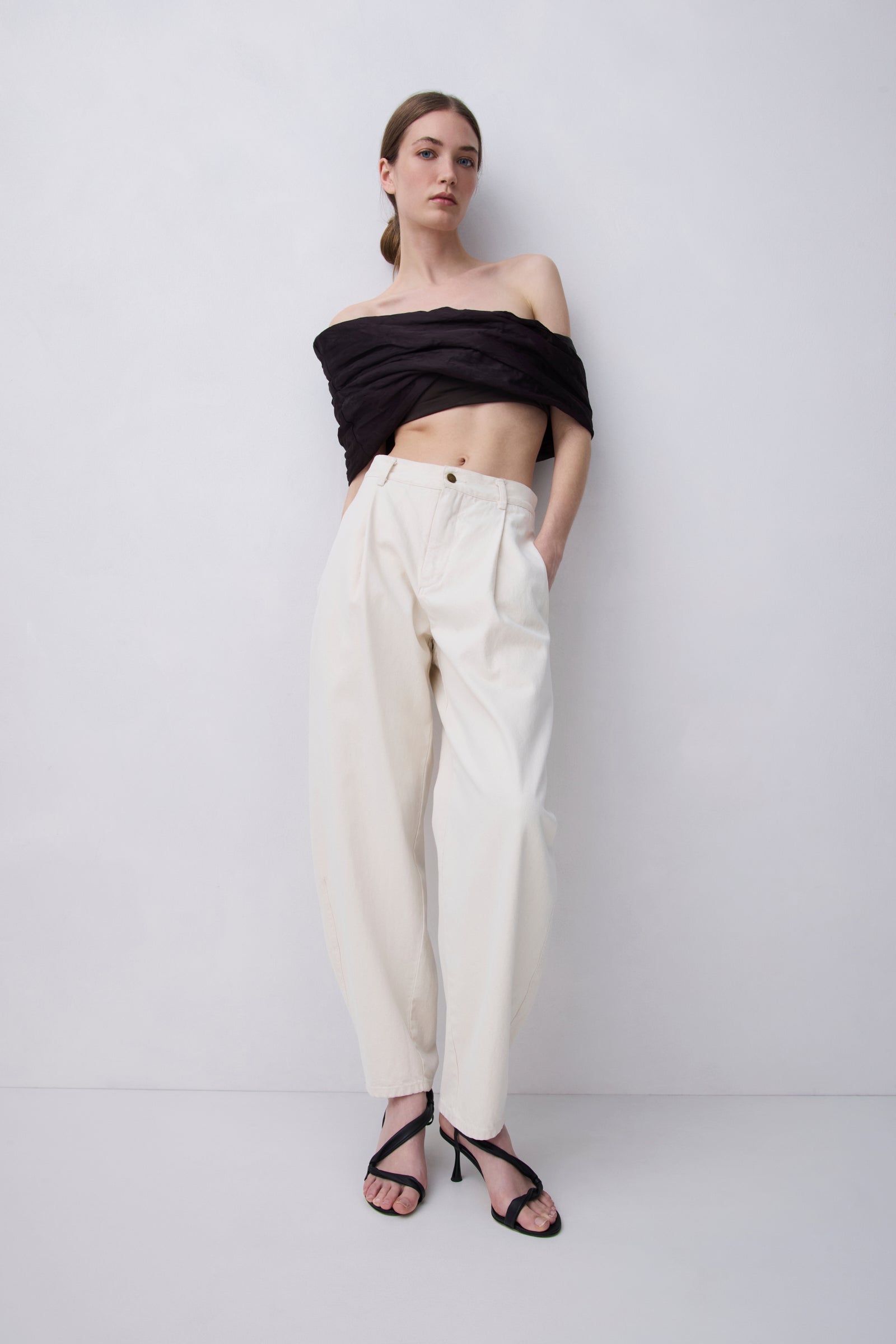 A model stands against a light gray studio wall wearing a black off-the-shoulder cropped top, cream wide-leg trousers, and black strappy high-heeled sandals.