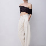 A model stands against a light gray studio background wearing a black off-the-shoulder wrap top and cream wide-leg trousers with black strappy sandals.