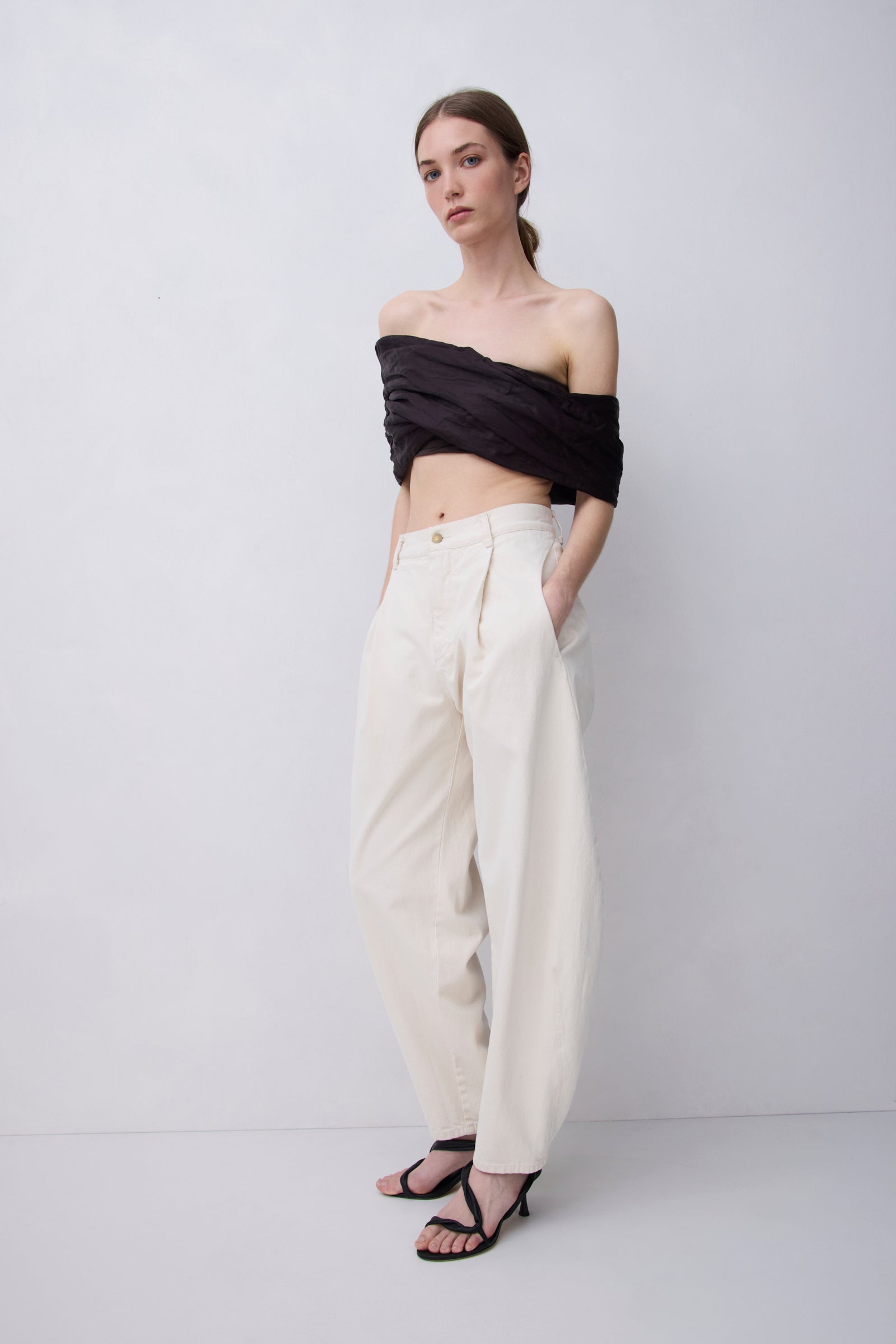 A model stands against a light gray studio background wearing a black off-the-shoulder wrap top and cream wide-leg trousers with black strappy sandals.