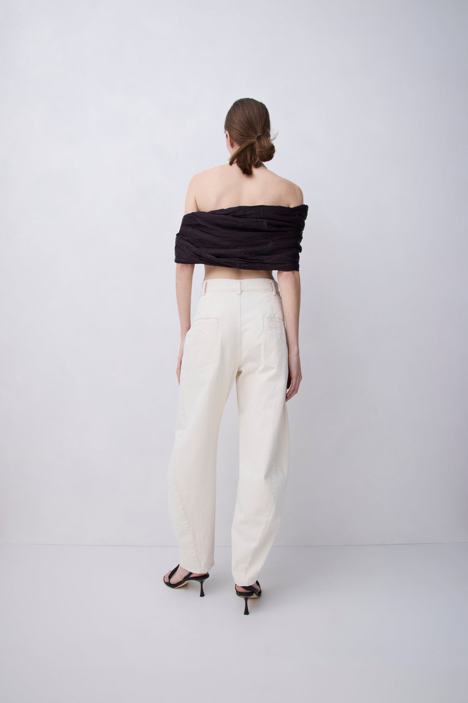 A model shown from the back wearing a black off-the-shoulder wrap top and cream wide-leg trousers with black strappy heels, set against a plain light gray studio background.