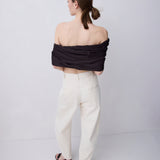 A woman is shown from behind wearing a black off-the-shoulder wrap top and cream-colored wide-leg trousers, standing against a plain light gray studio background.