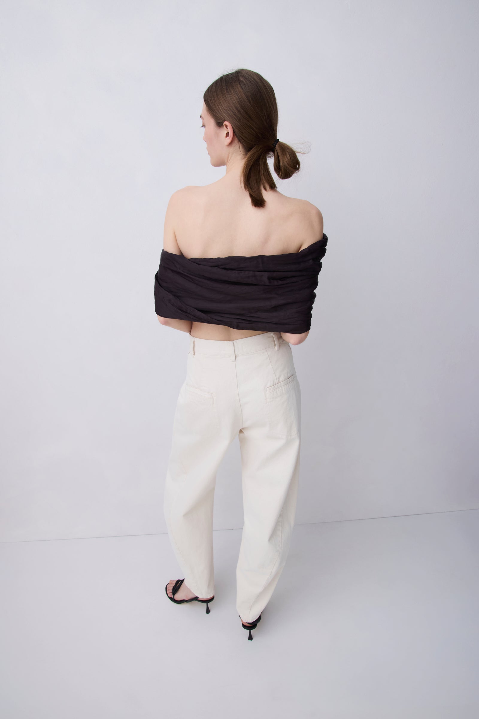 A woman is shown from behind wearing a black off-the-shoulder wrap top and cream-colored wide-leg trousers, standing against a plain light gray studio background.