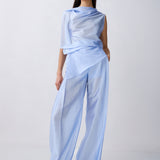 A model wearing a light blue draped, semi-sheer outfit consisting of an asymmetrical draped top and wide-leg trousers, standing against a plain light gray studio background.