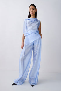 A model wearing a light blue draped, semi-sheer outfit consisting of an asymmetrical draped top and wide-leg trousers, standing against a plain light gray studio background.