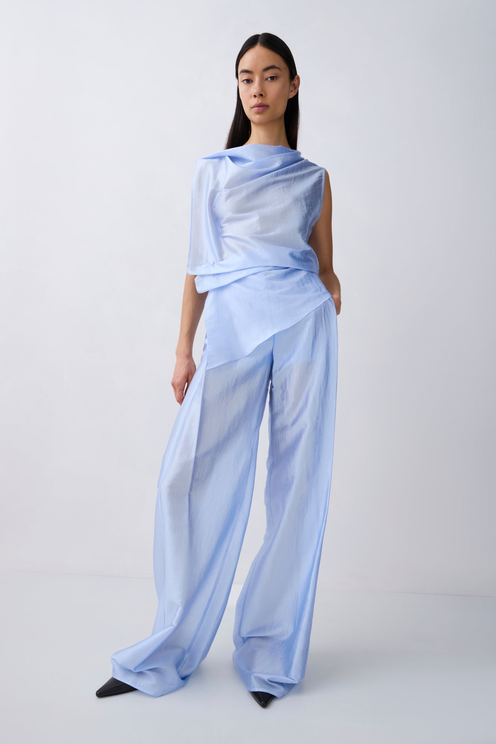 A model wearing a light blue draped, semi-sheer outfit consisting of an asymmetrical draped top and wide-leg trousers, standing against a plain light gray studio background.