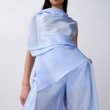 A model wearing a light blue draped top and matching wide-leg pants against a plain light gray studio background.