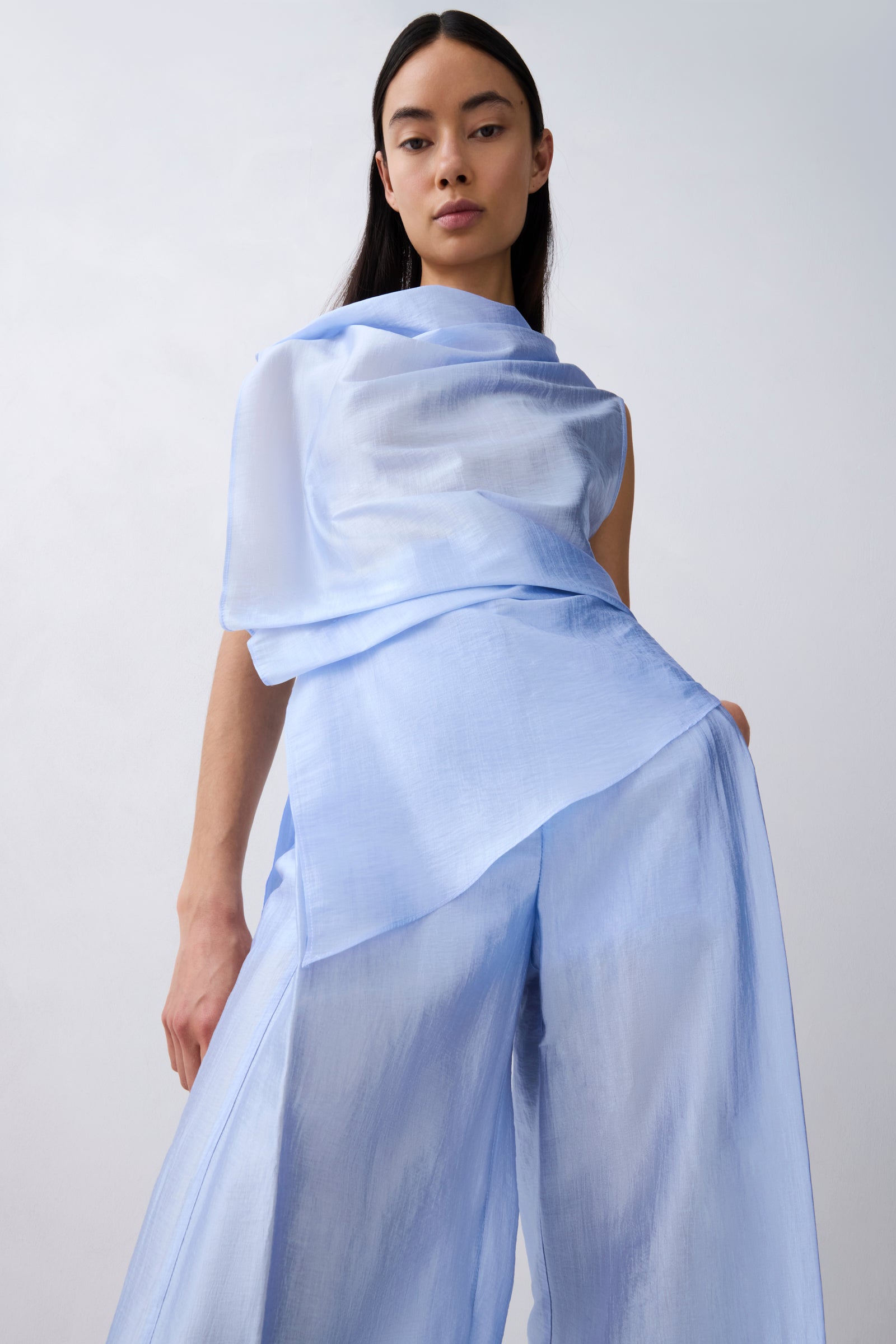 A model wearing a light blue draped top and matching wide-leg pants against a plain light gray studio background.