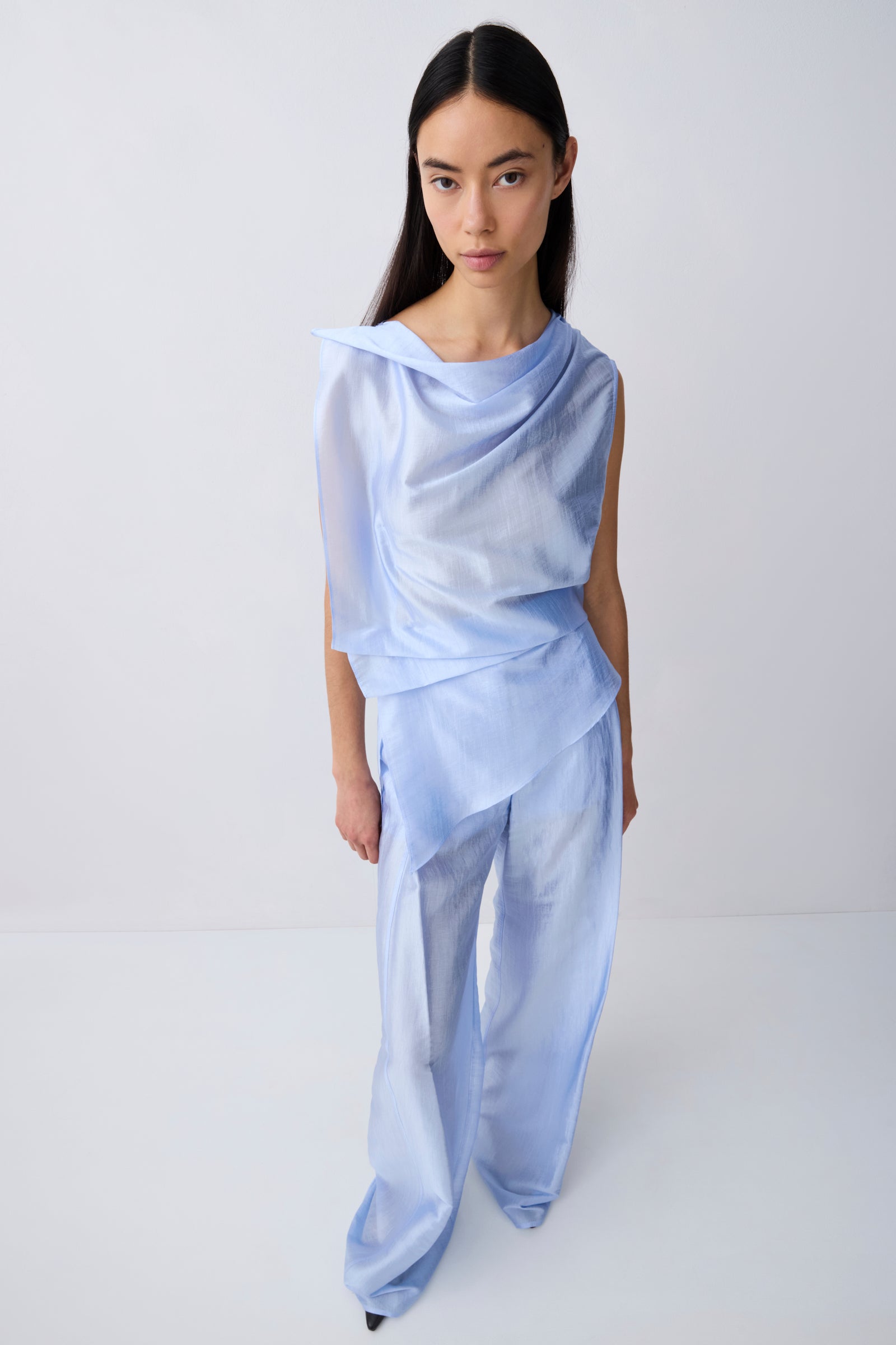 A model wearing a light blue draped two-piece outfit consisting of a sleeveless top and matching wide-leg pants, photographed against a seamless white studio background.