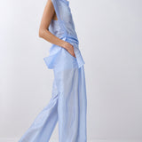 A model wearing a light blue sheer, draped two-piece outfit with wide-leg pants and a matching top, standing against a plain white studio background.