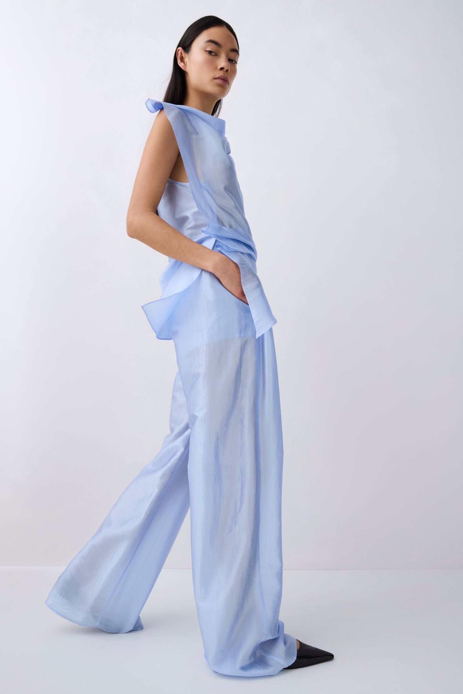 A model wearing a light blue sheer, draped two-piece outfit with wide-leg pants and a matching top, standing against a plain white studio background.