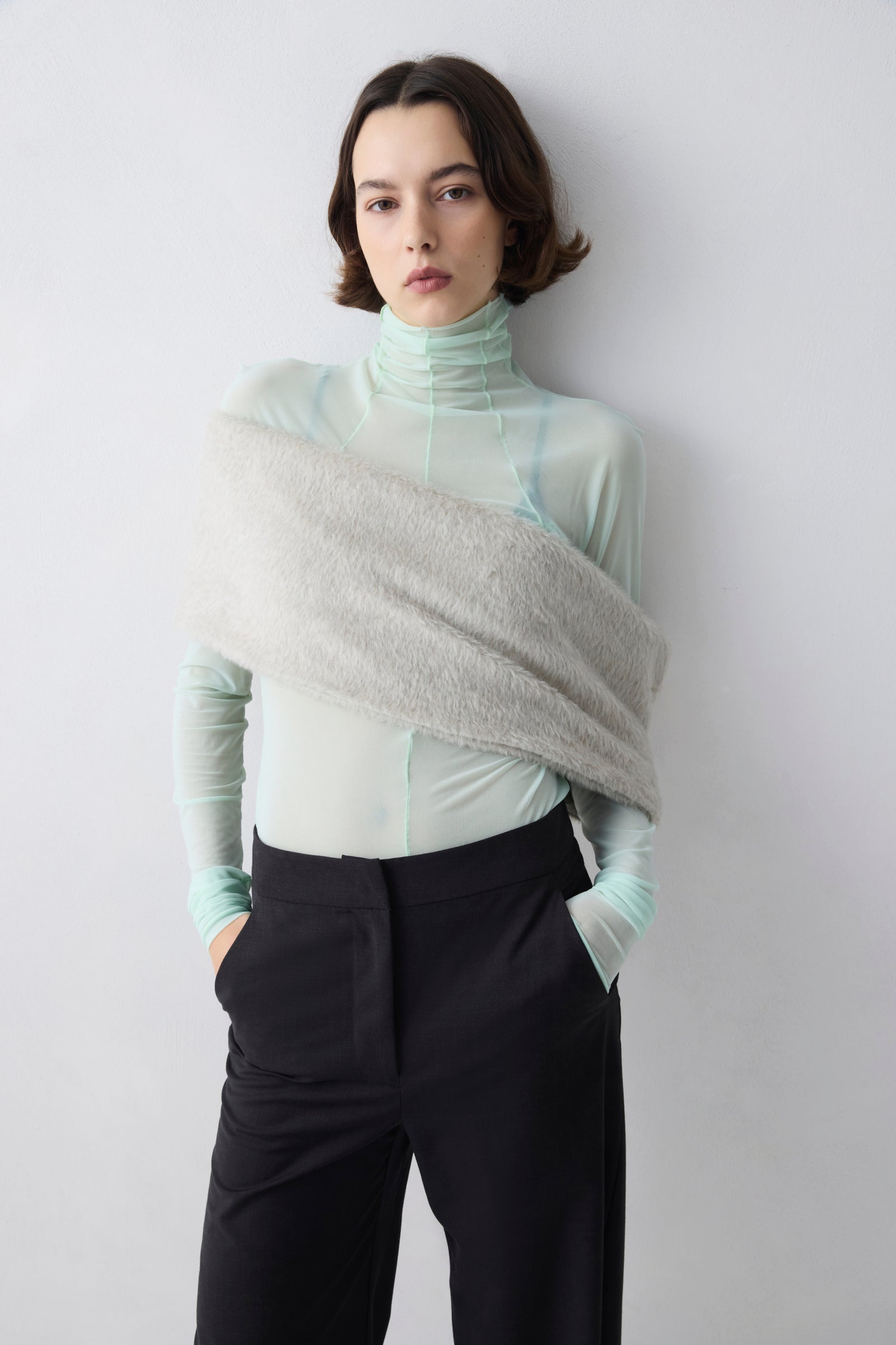 Person wearing a light green turtleneck and black pants with a gray shawl over one shoulder against a white background