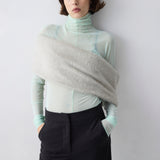 Person wearing a light green turtleneck and black pants with a gray shawl over one shoulder against a white background